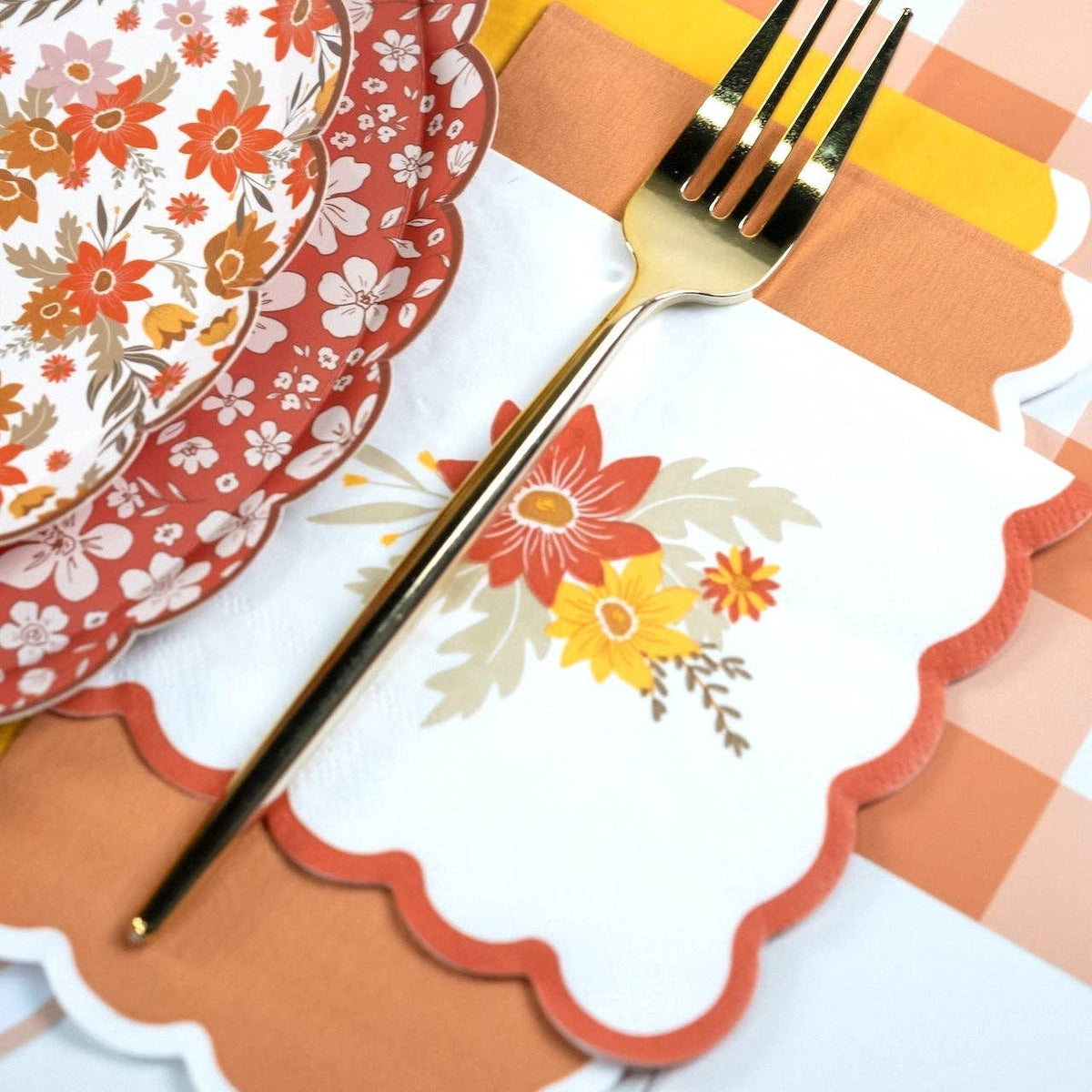Fall cocktail napkin under a gold fork in a place setting with floral paper plates and an orange gingham runner