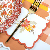 Fall cocktail napkin under a gold fork in a place setting with floral paper plates and an orange gingham runner