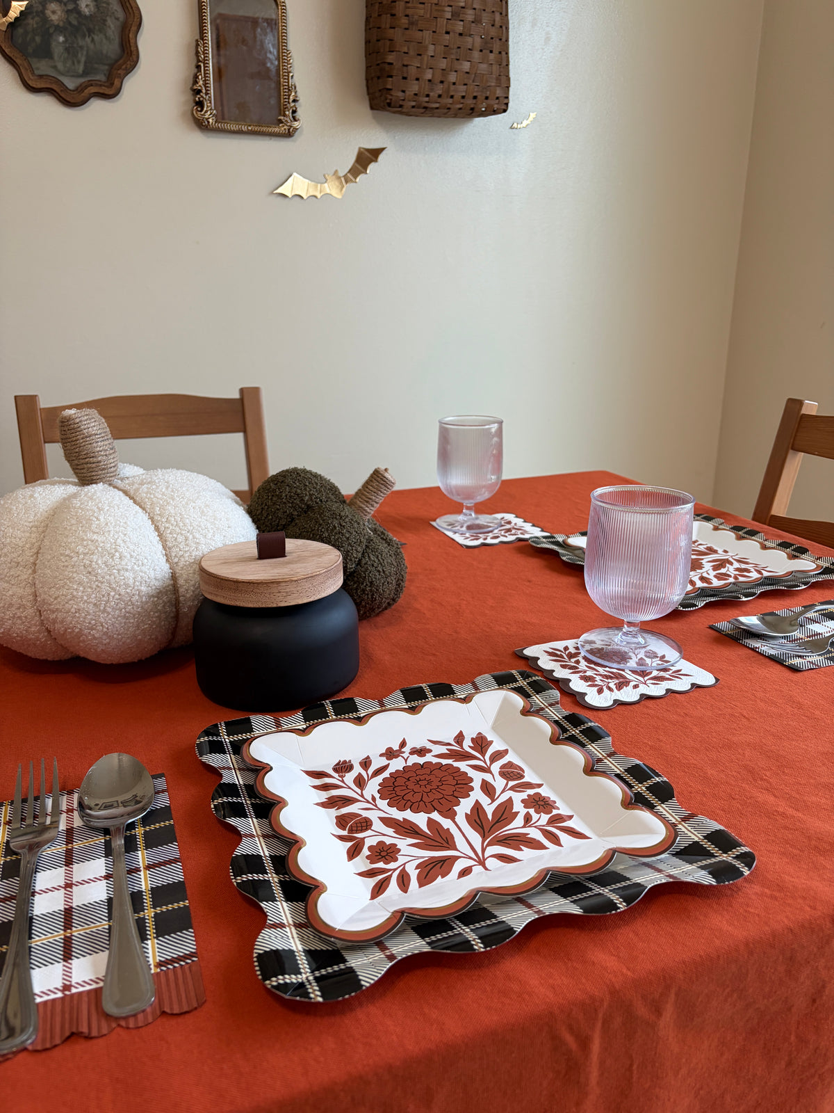 Fall dining room table styled with rust floral square paper plates, knit pumpkins, black and white plaid placemats, pink glasses, and wood chairs