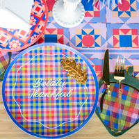 Overhead place setting showing the acorn napkin tucked on a plaid plate with a wheat sprig, gold flatware, and a geometric-patterned plate.