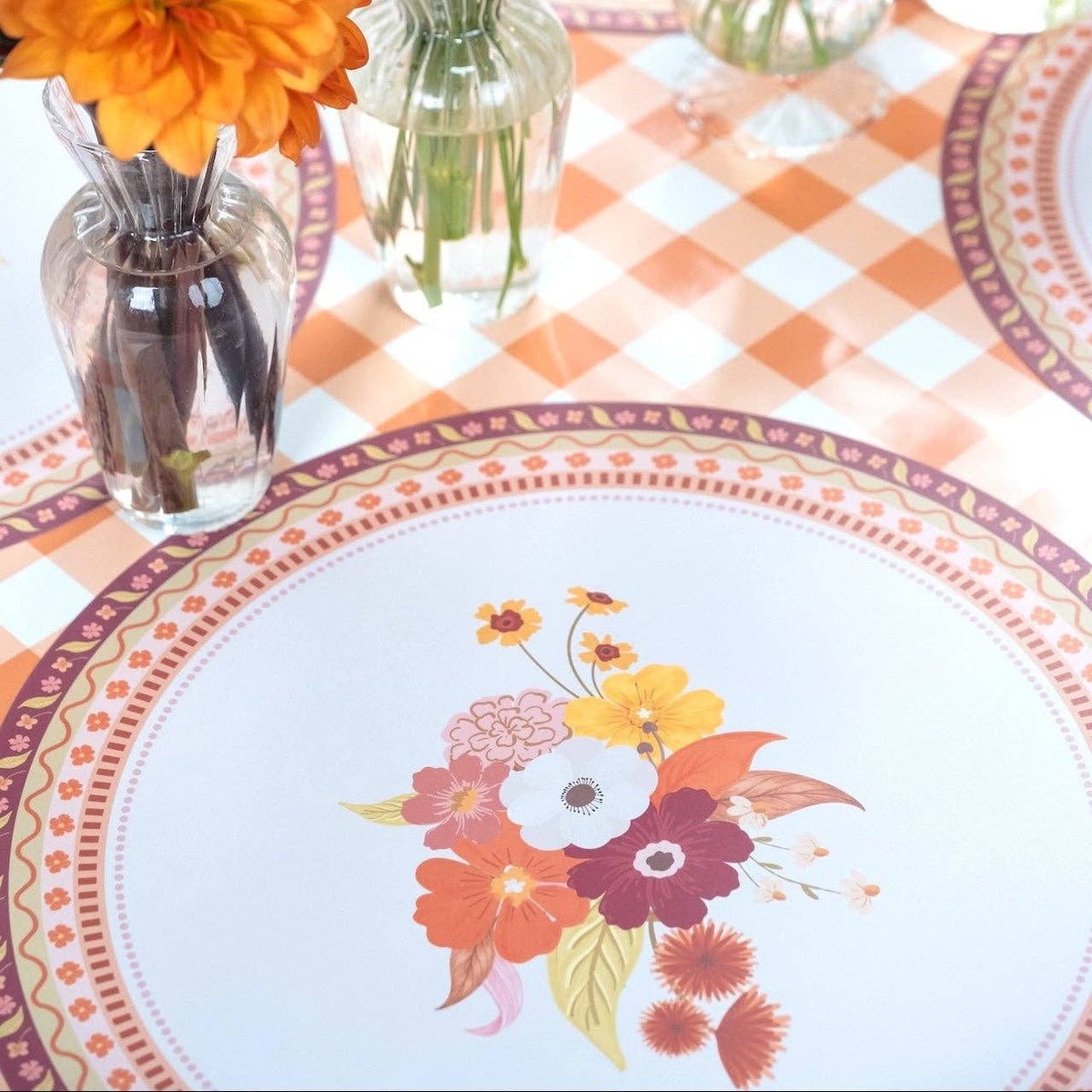 Styled fall place setting showing the floral placemat as the base under a white floral plate, with a black cat figure and gingham runner.