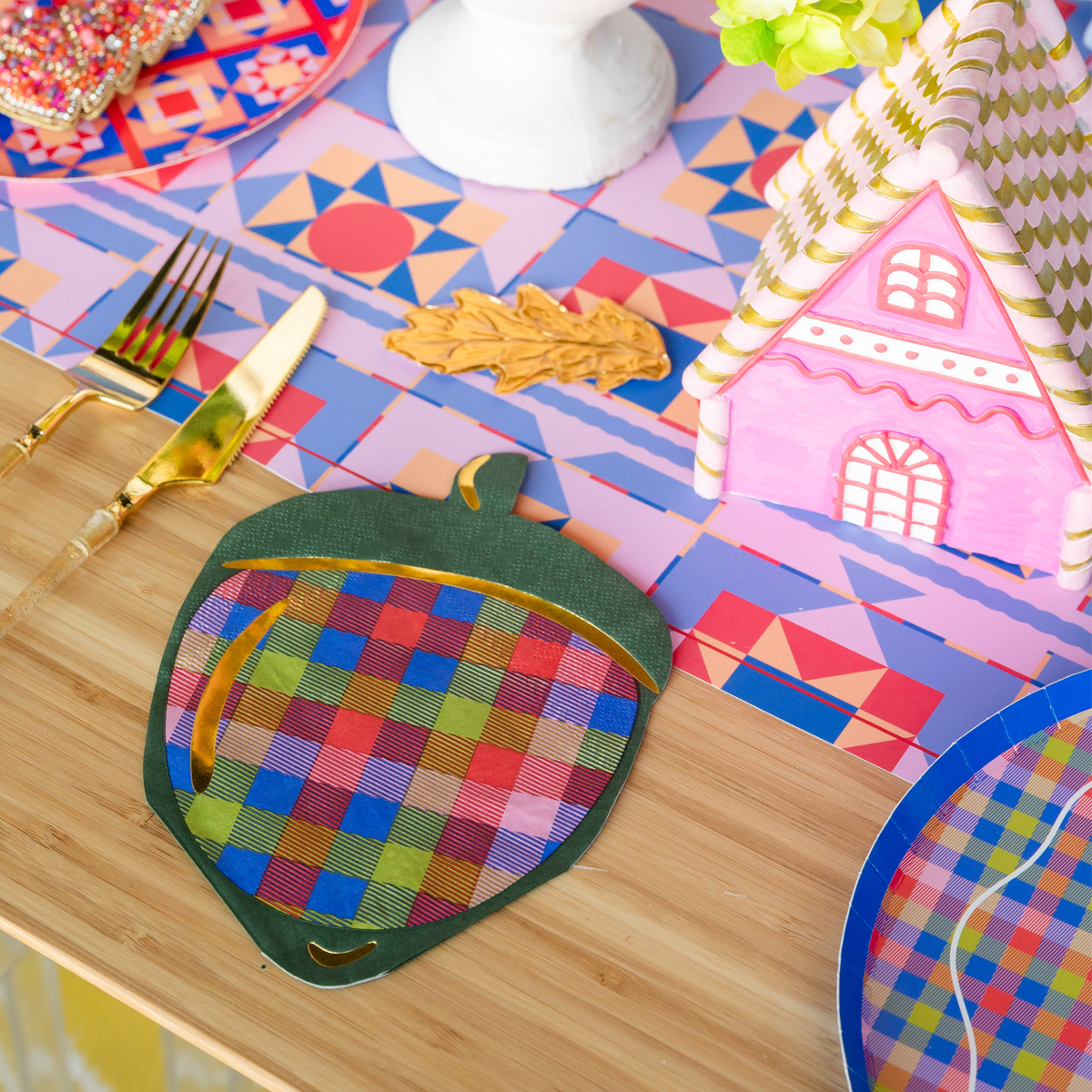 Overhead fall place setting showing the plaid acorn napkin beside a plate with a gingerbread house and colorful autumn décor on a plaid runner.