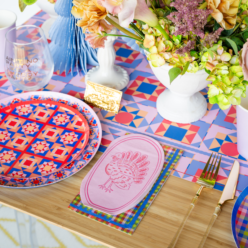 Overhead fall place setting showing the purple turkey napkin with geometric-patterned plates, gold flatware, and autumn florals on a plaid runner.