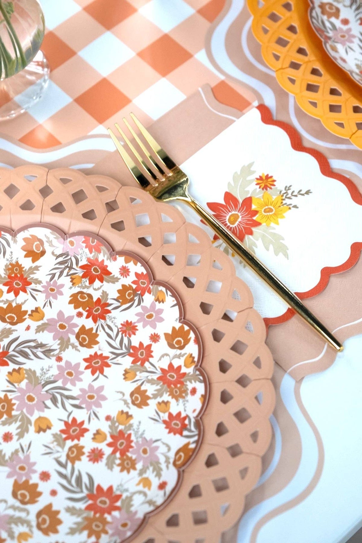Fall place setting close-up showing the tan napkin with a floral plate, gold flatware, and a leaf-print napkin on a gingham runner.