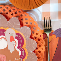 Close-up fall place setting showing the turkey plate on an orange lattice charger with a burgundy napkin and gold fork.