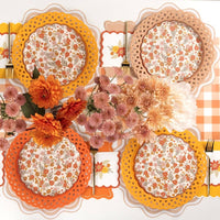 Styled fall table overhead — orange floral plates on scalloped chargers, pink and orange flowers, orange napkins, and orange-and-white gingham runner.