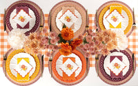 Overhead fall tablescape — orange floral plates, cream chargers, orange napkins, and a warm autumn spread on a gingham runner.
