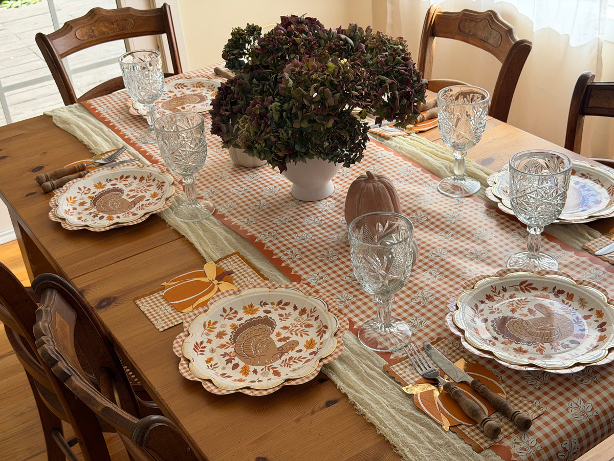 Round fall turkey paper plates with gold scalloped edges layered on gingham plates along an orange gingham floral table runner with crystal goblets and pumpkins