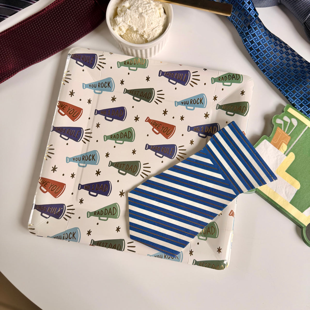 Overhead flatlay of a megaphone print paper dinner plate topped with a blue striped tie-shaped napkin, golf cart napkin at right, cream cheese ramekin and gold butter knife above, neckties used as styling props on a white round table