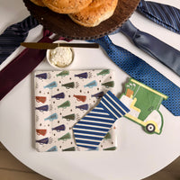 Flatlay of Father's Day party tableware: megaphone print square paper dinner plate centered on a white table with a blue striped tie-shaped cocktail napkin and golf cart shaped napkin, surrounded by neckties and a bagel board
