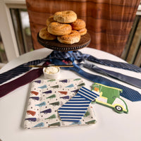 close-up flatlay of a megaphone print square paper plate printed with "Rad Dad," "Best Dad," "You Rock," and "Love You" text, layered with a blue and white striped tie-shaped napkin and golf cart napkin, neckties and cream cheese ramekin in the background