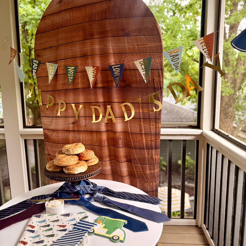 Father's Day party table styled against a wood-grain arch backdrop with a gold "Happy Dad's Day" letter banner and multicolor pennant garland above, bagels on a cake stand, and megaphone plates, striped tie napkins, and golf cart napkin on the table below
