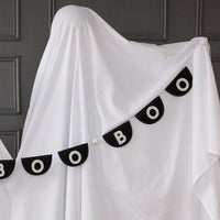 Cinemagraph-style panorama of Boo felt Halloween banner strung in a gentle smile between two sheet ghosts. Slate gray paneled wall and rustic brick create modern-farmhouse setting; banner’s matte black felt contrasts sharply against soft draped fabric. White stitched letters and mini pom-poms pop for high visibility in party photos, showcasing how the six-foot garland fills wider spaces like mantels, stair rails or porch beams with minimal effort.