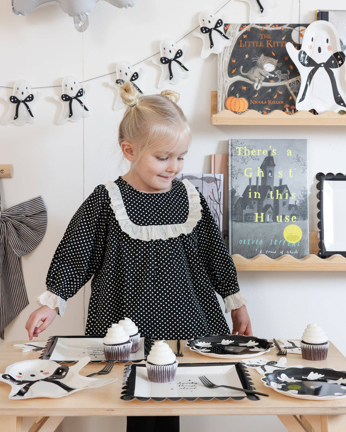 Child in a polka dot dress standing beside a table with ghost felt Halloween-themed decor.