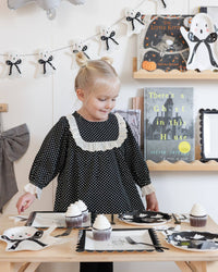 Child in a polka dot dress standing beside a table with ghost felt Halloween-themed decor.