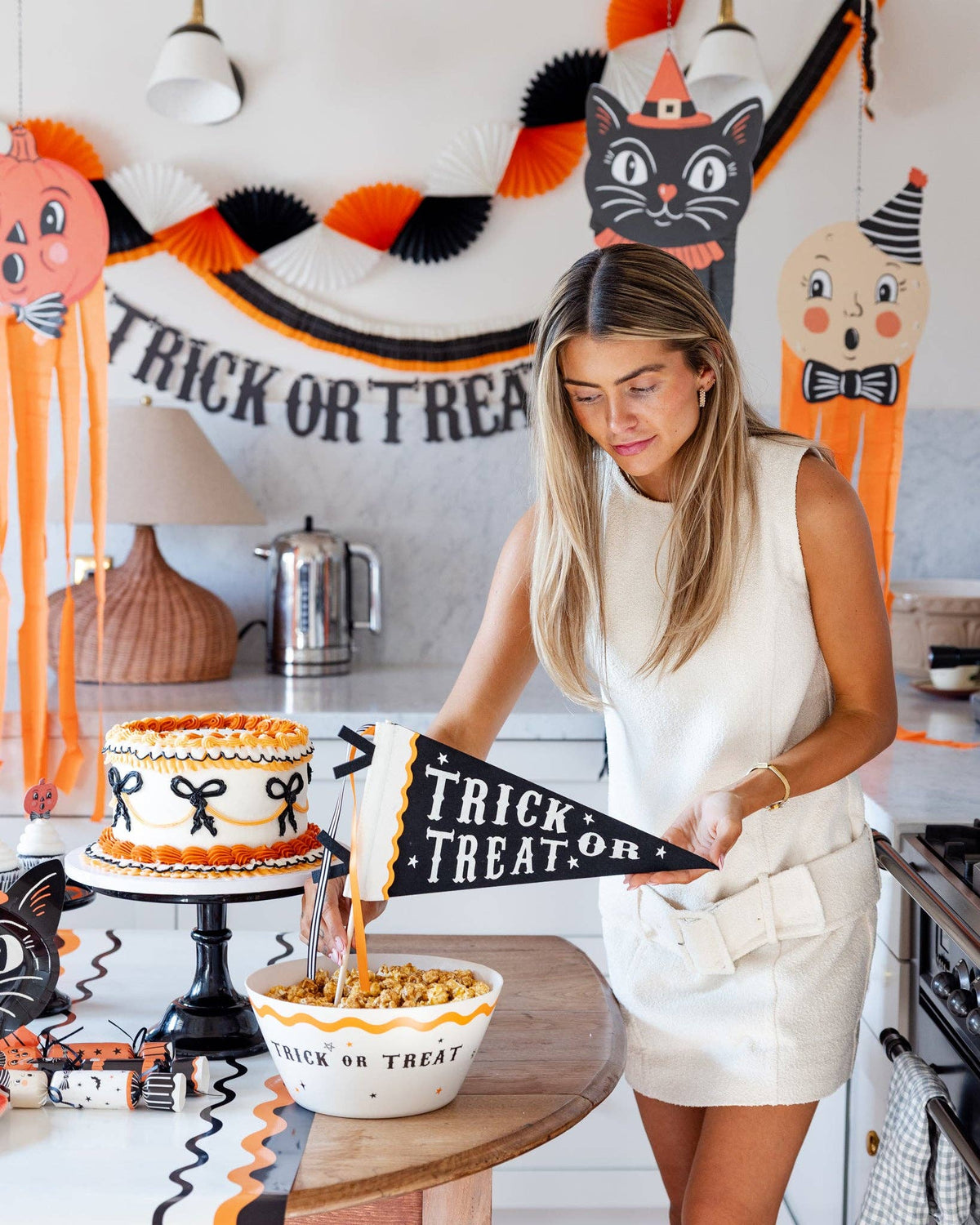 Woman in a kitchen with Halloween decorations and a cake with the Trick or Treat Halloween felt pennant.