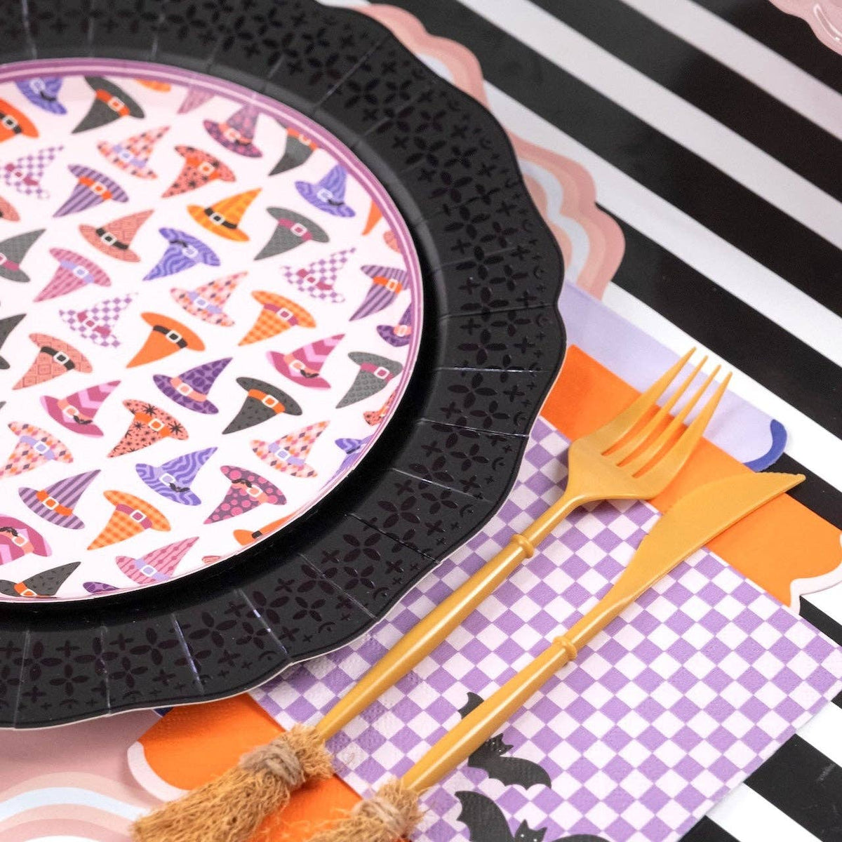 Styled feminine Halloween place setting — witch hat plate on a black textured scalloped charger with a purple checkered napkin and gold flatware on a black-and-white striped table.