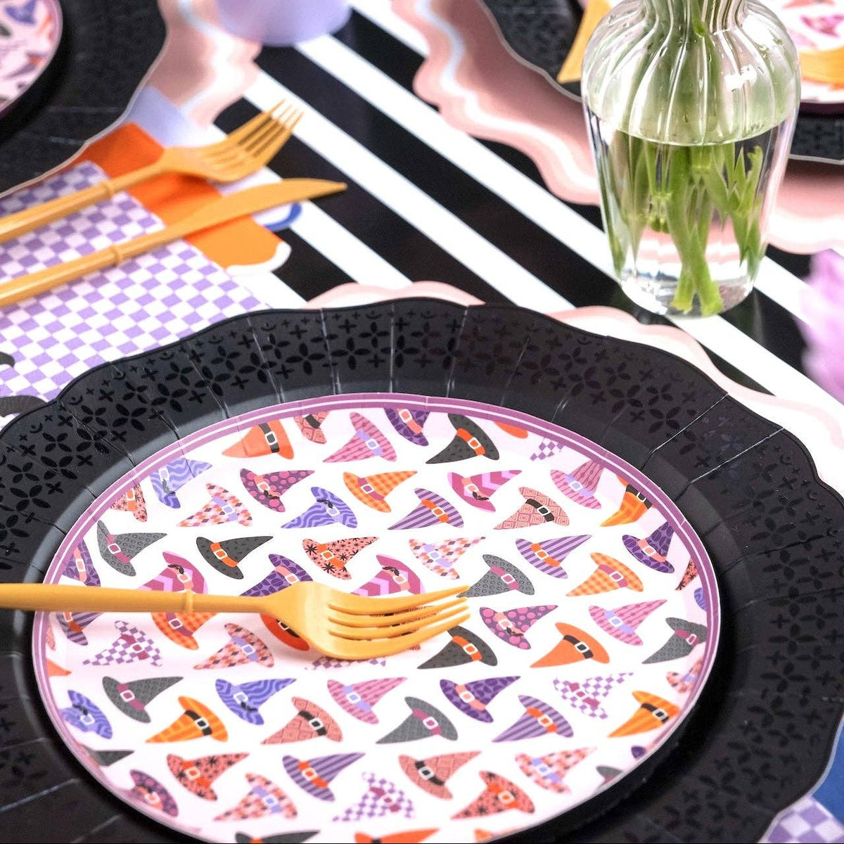 Angled view of the feminine Halloween table showing witch hat plates, pink roses in glass vases, purple checkered details, and gold flatware from a seated perspective.