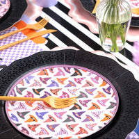 Angled view of the feminine Halloween table showing witch hat plates, pink roses in glass vases, purple checkered details, and gold flatware from a seated perspective.