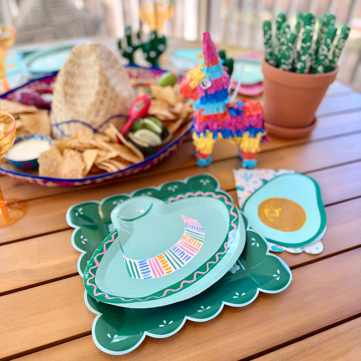 Fiesta place setting with green papel picado dinner plate and sombrero-shaped plate alongside avocado napkin, mini piñata, and cactus straws on a wood table