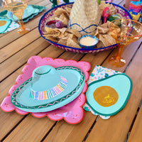 Close-up fiesta place setting with pink papel picado dinner plate layered with sombrero-shaped plate, avocado-shaped napkin, and cacti print napkin beside a margarita glass