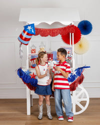 Two children standing in front of a 4th of July party cart decorated with fringe banners and paper fans, with a firecracker shaped foil balloon attached to the top left corner