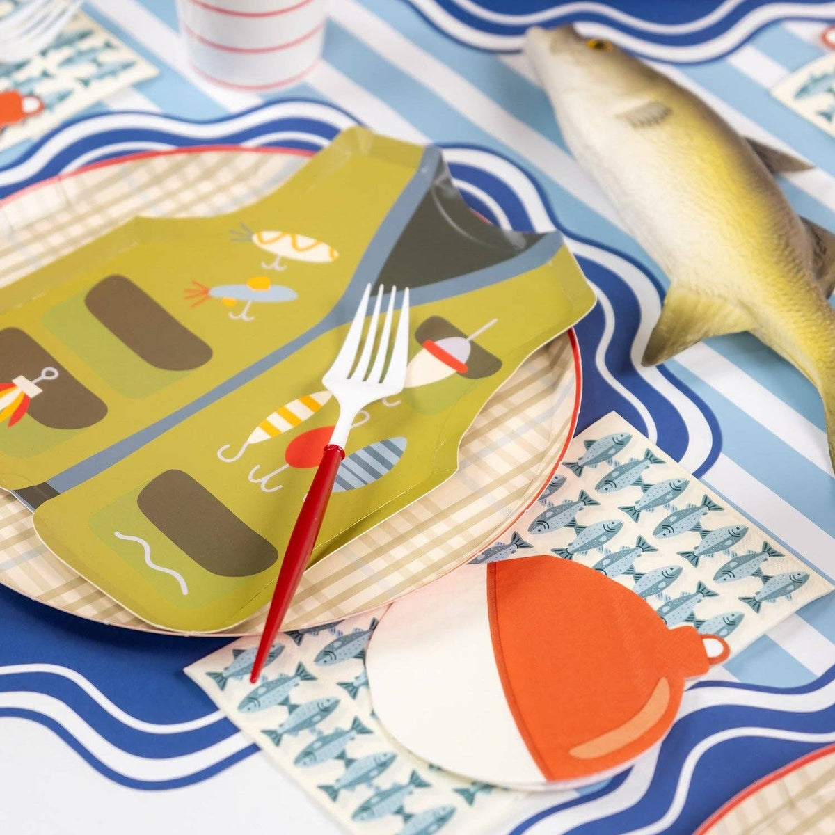 Fishing vest plate on a plaid plate with striped cups and a fishnet on a wavy blue placemat.