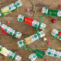 Overhead flat lay of several Christmas crackers scattered on a wooden table with greenery, ready for use as Christmas dinner favors.