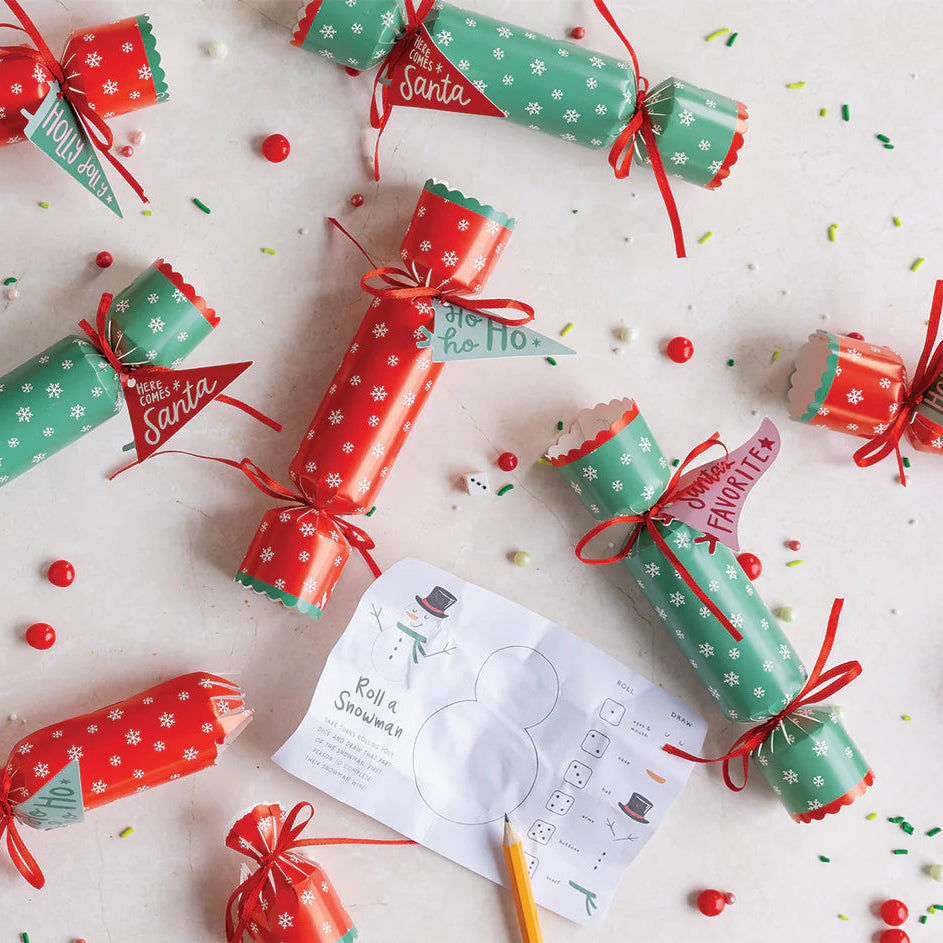 Overhead flat lay showing multiple holiday poppers scattered with the "Roll a Snowman" interactive dinner game card.