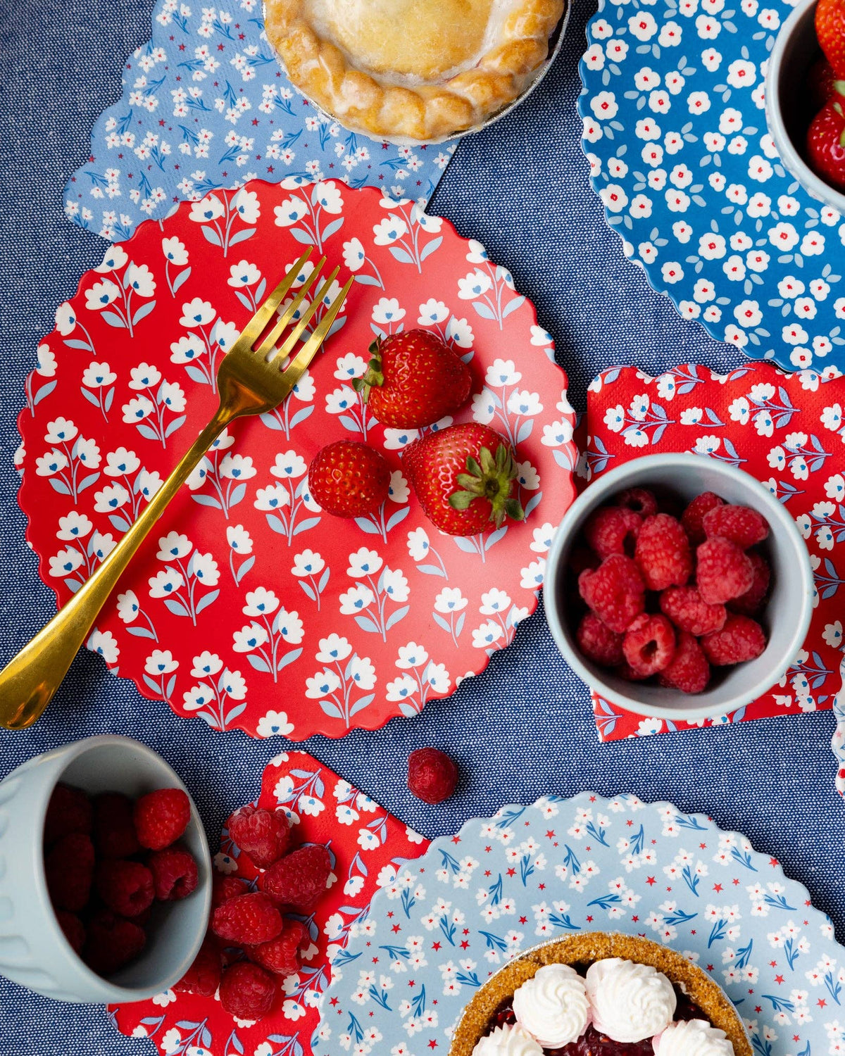 Overhead table setting with all four floral bamboo plates layered on blue tablecloth with strawberries, raspberries, pie, and gold forks
