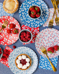 Overhead styled table with all four floral bamboo plate designs, fresh berries, pies with whipped cream, and gold forks on blue tablecloth