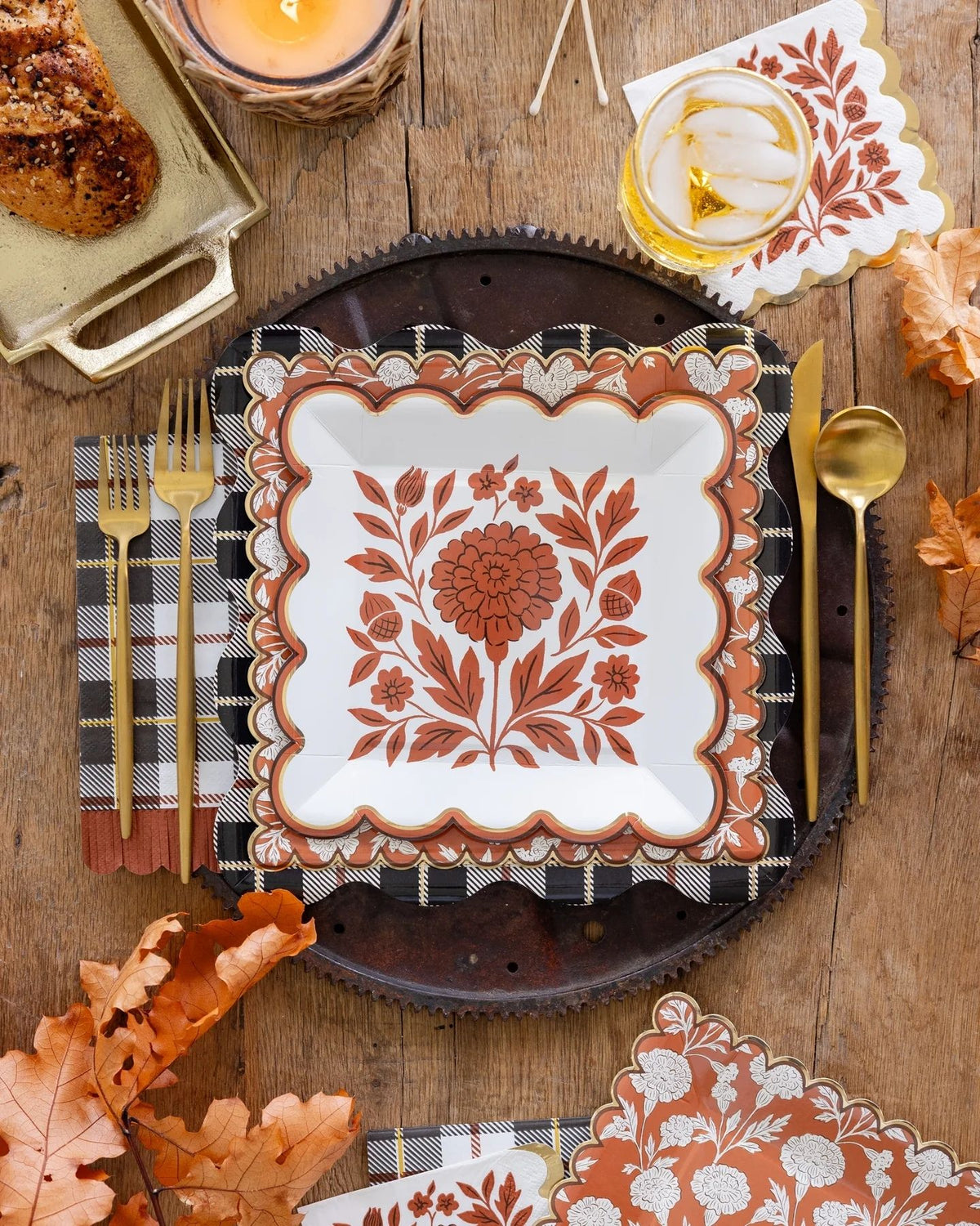 A styled autumn table setting featuring a white square plate with a terracotta floral design, a black-and-white gingham napkin with gold flatware, matching patterned paper napkins, and fall leaves on a rustic wooden table.