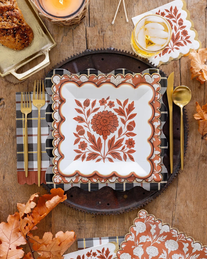 A styled autumn table setting featuring a white square plate with a terracotta floral design, a black-and-white gingham napkin with gold flatware, matching patterned paper napkins, and fall leaves on a rustic wooden table.