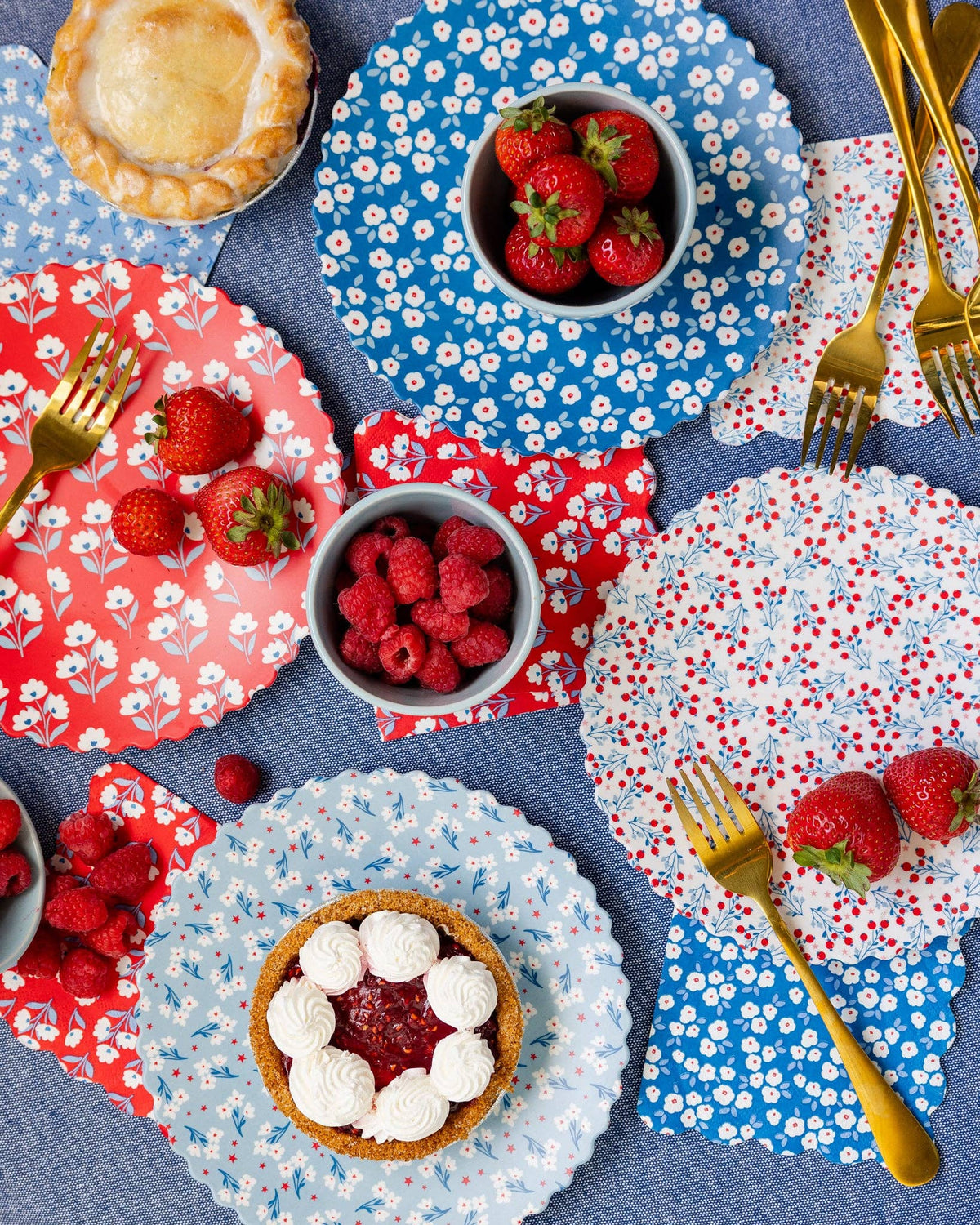 Overhead styled table with all four floral napkin and bamboo plate designs layered with strawberries, raspberries, pie, and gold forks on blue tablecloth