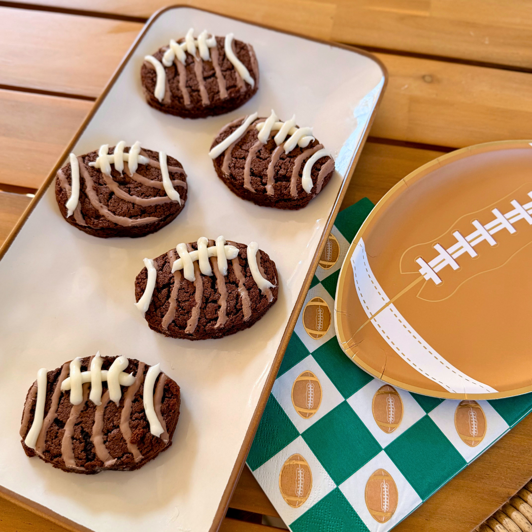 A white rectangular serving platter filled with round chocolate cookies decorated with white icing laces to look like footballs, set on a wooden table.
