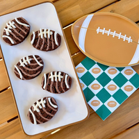 A top-down view of a game day party spread on a wooden table, featuring a white platter of football-decorated cookies next to a football-shaped plate and patterned napkins.