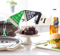 Three mini football pennants staked in stacked brownies on a white cake stand at a game day spread