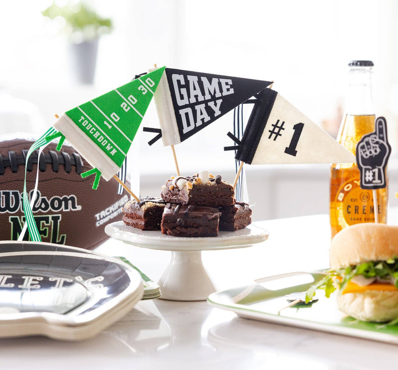 Three mini football pennants staked in stacked brownies on a white cake stand at a game day spread