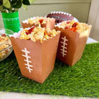 A close-up photograph of four novelty football-themed popcorn boxes placed on artificial turf. The brown paper boxes, with white painted laces, are filled with savory popcorn featuring red pepperoni slices and cheese. In the background, there is an authentic leather American football with 'Wilson' branding and white laces, next to a green vase with eucalyptus sprigs, all set against a sage-green wall.