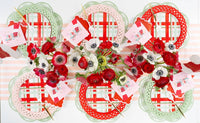Symmetrical overhead flat lay of four holiday place settings arranged in a line with red scalloped chargers, plaid plates, and pink cocktail napkins.
