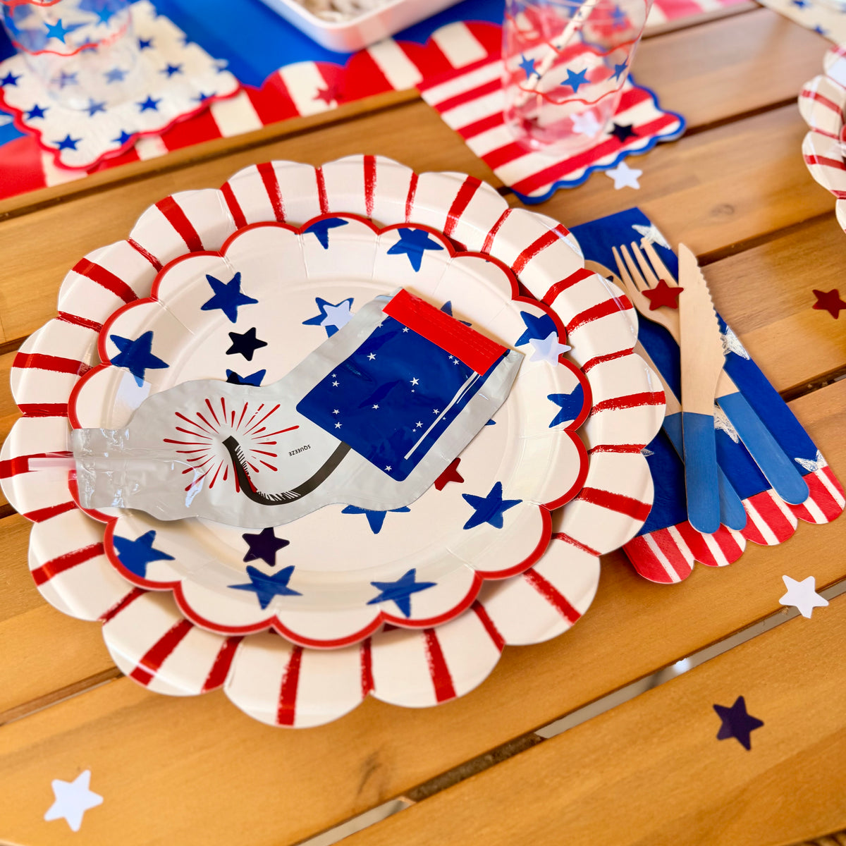 Fourth of July party plates with red stripes and blue stars, paired with matching napkins and clear cups on a wooden table.