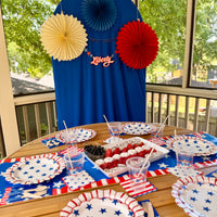 Fourth of July party table setup with matching plates, cups, and a red, white, and blue backdrop with paper fans and banner.