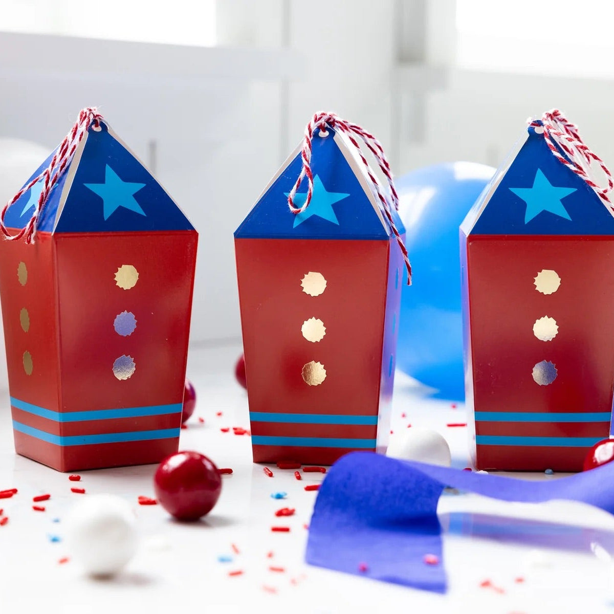 Set of three red, white, and blue rocket-themed treat boxes arranged with party decorations and sweets.