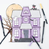 Overhead view of a fully colored haunted house paper placemat surrounded by scattered pastel markers.