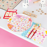 Game night round paper dinner plate on a blue checkered napkin styled with a Scrabble board, white dice decor, jackpot slot machine shaped plate, gold cutlery, and game night cocktail napkins