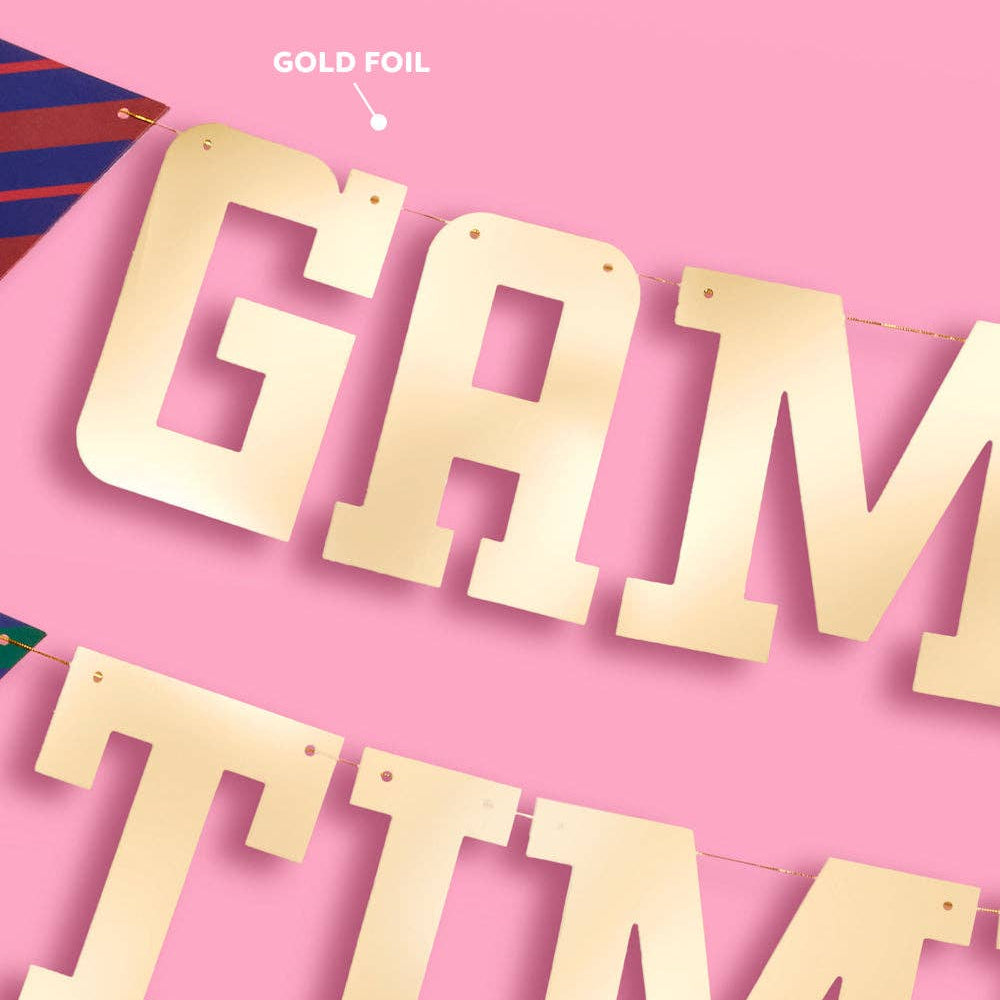 Close-up of gold foil “GAME TIME” letter banner with pre-strung holes on pink background