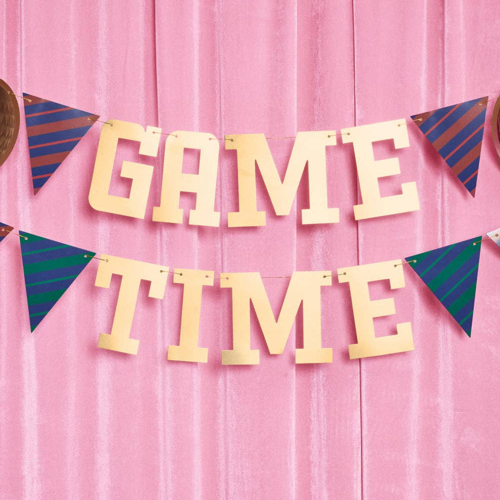 Gold foil “Game Time” banner with striped pennants hanging on a pink backdrop