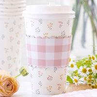 A lifestyle photo of a floral paper coffee cup with a pink gingham sleeve sitting on a marble table next to spring flowers and ranunculus.