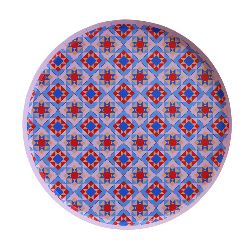 Round geometric holiday paper plate with an all-over folk-art medallion pattern in blue, red, and pink, overhead view on white background.