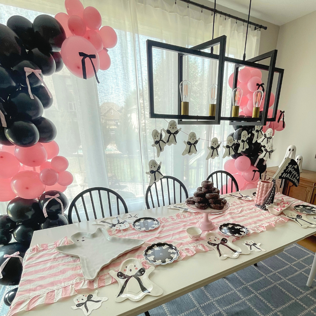 A styled Pink and black Halloween party tablescape in a room with black and pink balloon garlands, ghost decorations, a pink-and-white striped table runner, plates, and napkins.